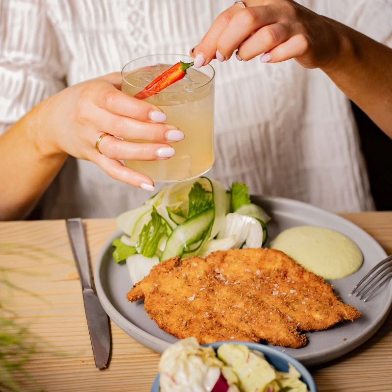 Woman holding a spicy margarita with a red chili above a plate of chicken Milanese with seasonal greens