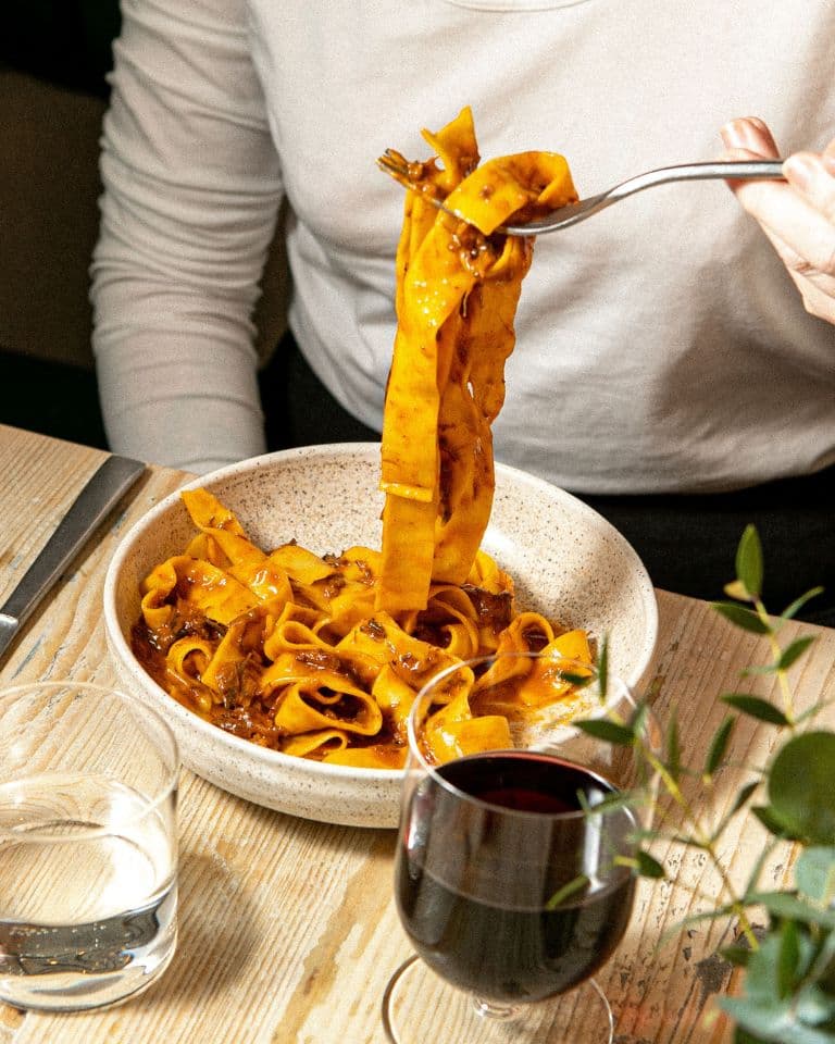 bowl of beef shin ragu pasta alongside a glass of red wine and water